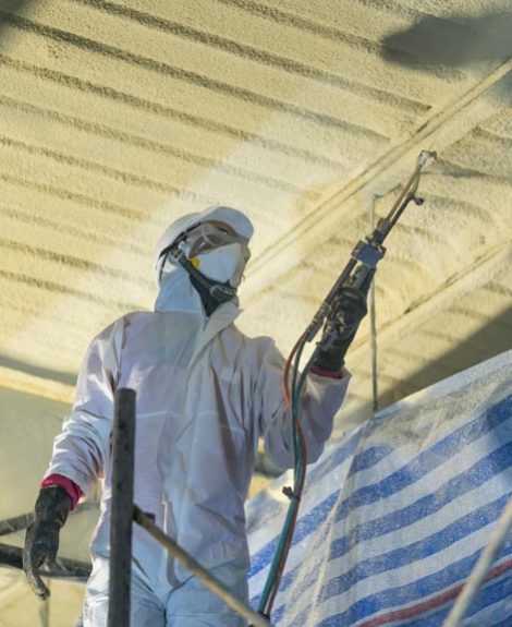 A professional in full gear and in action of applying a spray foam roofing system which can help improve a roof’s structural integrity by locking its components in place